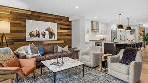 A modern living room with a brown leather sofa, a grey armchair, and a coffee table. There is a wooden wall with a bear painting and a kitchen in the background. - Luxury Escape | Private Hot Tub and Firepit (Canmore)