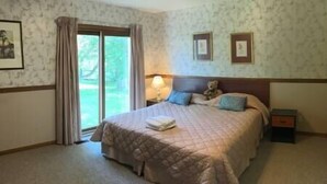Un dormitorio con una cama grande, una mesita de noche, una lámpara y un osito de peluche. Se ve una ventana con cortinas y una puerta que da al exterior.