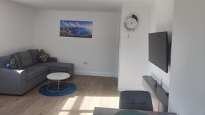 Una sala moderna con un sofá seccional gris, una mesa de centro redonda, un cuadro de un paisaje costero, un reloj de pared y un televisor de pantalla plana.