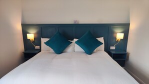 Una cama bien tendida con sábanas blancas y dos almohadas color turquesa, flanqueada por mesitas de noche con lámparas de pared.