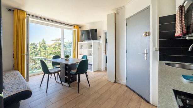 A compact living space with a balcony, a dining area, a kitchenette, and a TV mounted on the wall. - Le Crépuscule - Wifi - Balcon (Créteil)