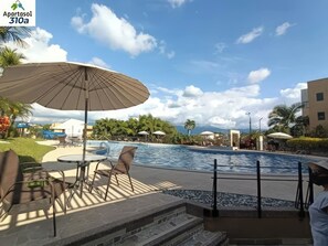 Piscina com espreguiçadeiras e guarda-sóis, cercada por vegeta ção e edifícios.