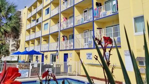 A yellow multi-story hotel with balconies, a swimming pool, and palm trees.