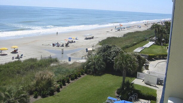 A beach view with umbrellas, lounge chairs, and a grassy area with a fence.