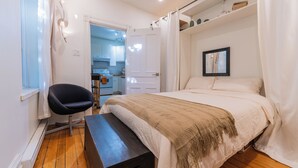 Une chambre à coucher comprenant un lit, une chaise, un banc, une fenêtre et une porte donnant sur la cuisine.