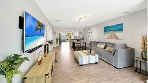 Un salon moderne avec un téléviseur à écran plat, un canapé gris, un pouf rayé et une table basse en bois. Deux tableaux ornent les murs, ainsi qu’un banc en bois et une plante en pot.
