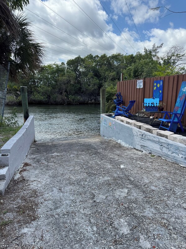 A concrete pathway leading to a dock with blue chairs and a table, surrounded by a wooden fence and greenery.