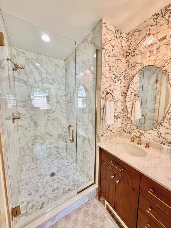Un baño moderno con ducha envuelta en vidrio, paredes de mármol y tocador con un espejo grande.