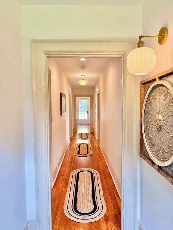 A hallway with wooden flooring, white walls, and a decorative wall piece. There are three rugs on the floor and a light fixture on the wall.