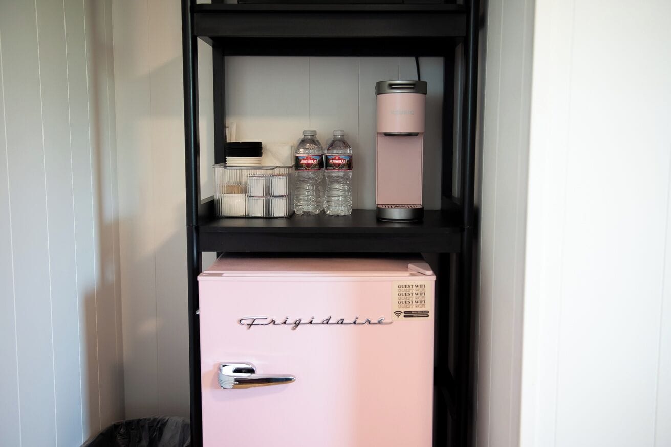 A pink minibar with a 'Frigidaire' label, two water bottles, and a pink thermos on a black shelf.