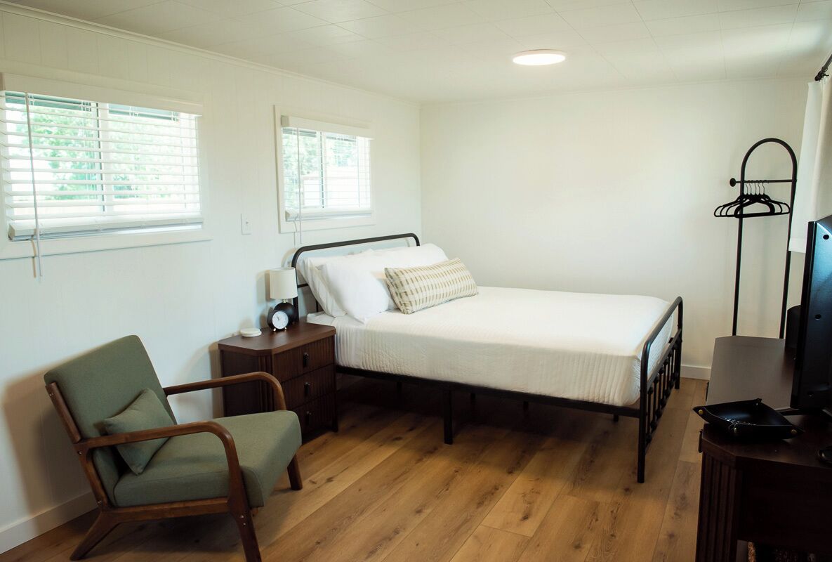A bedroom with a single bed, a chair, a bedside table, a lamp, and a TV stand.