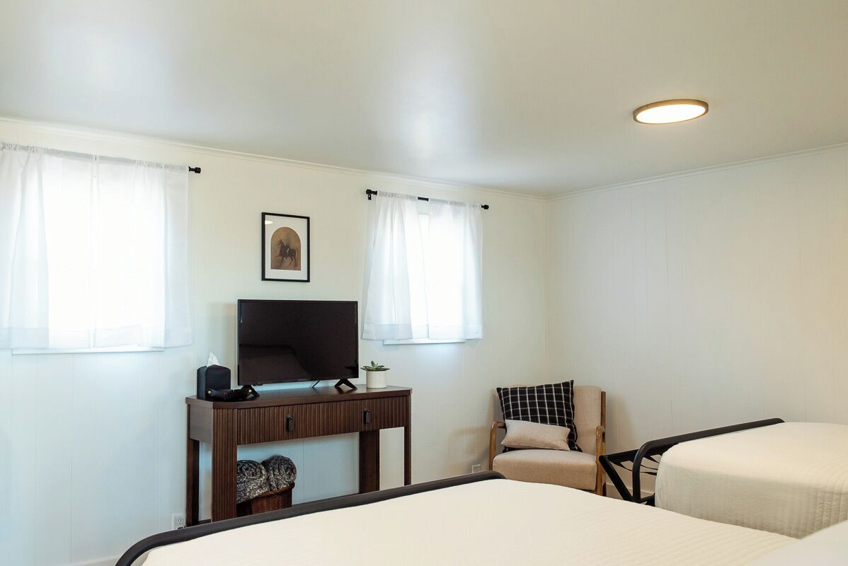 Ett hotellrum med en säng, ett skrivbord med en tv, en stol och två fönster med gardiner.