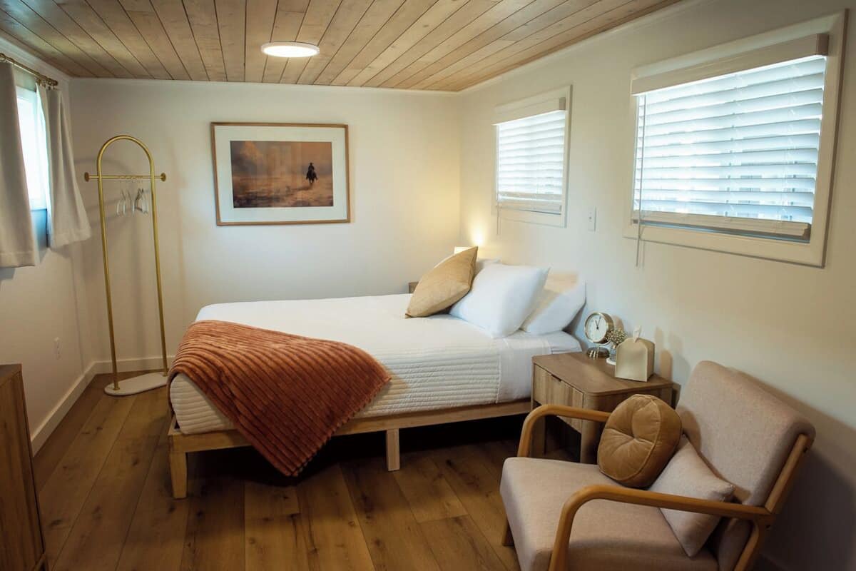 A bedroom with a bed, a chair, a bedside table with a clock, and a picture on the wall.
