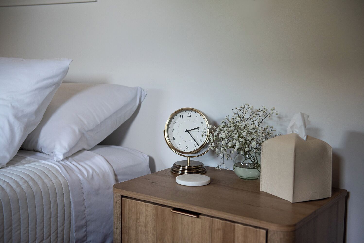 A neatly made bed with white linens, a bedside table with a clock, a vase with flowers, and a tissue box.