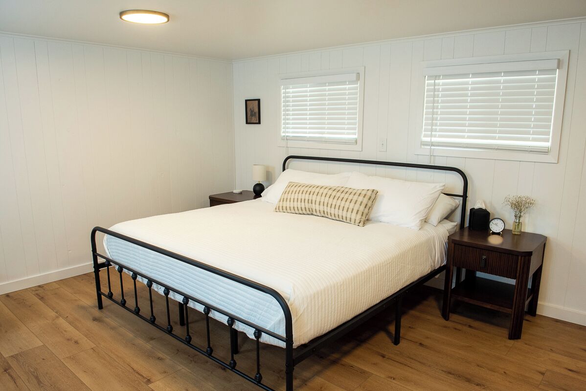 A bedroom with a metal bed frame, white bedding, two bedside tables with lamps, and a window with blinds.