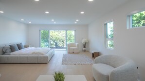 A modern bedroom with a large bed, a white armchair, and a view of greenery through the windows.