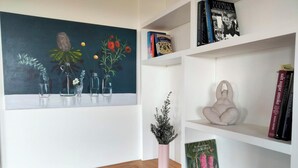 A room with a wooden desk, a white shelf with books, a vase with flowers, and a painting of flowers on the wall.