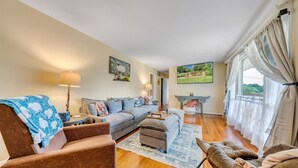 Una sala de estar con un sofá, sillones, una mesa de centro y un televisor. Hay una chimenea y una ventana con cortinas.