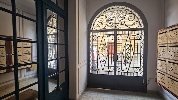 A room with a large arched window with intricate metalwork, a black door with glass panels, and a tiled floor. - Studio Cozy Ivry near Paris (Ivry-sur-Seine)