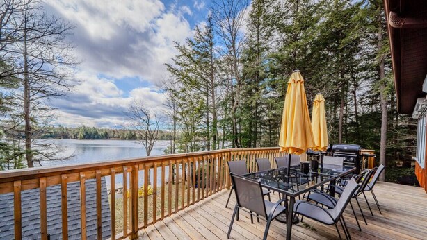 A wooden deck with a glass table and chairs, overlooking a lake with trees.