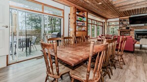 A spacious room with a large wooden dining table surrounded by chairs, a bookshelf, a fireplace, and large windows offering a view of the outdoors.