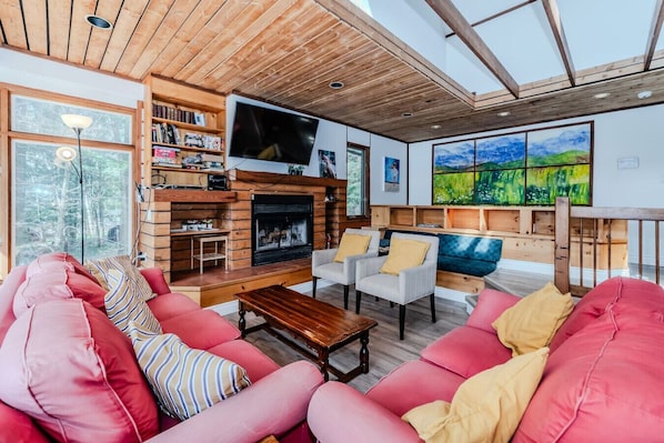 A cozy living room with a fireplace, wooden bookshelves, a flat-screen TV, and a large window overlooking a forest.