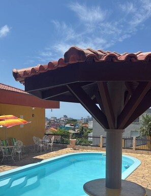 Área de piscina residencial con estructura de techo azulejado, mobiliario exterior y vistas a la ciudad al fondo.