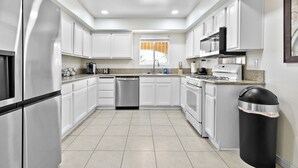 A modern kitchen with white cabinets, stainless steel appliances, and a black trash bin. - Patio w/ Grill + Pool Table , In-Unit Laundry (North Las Vegas)