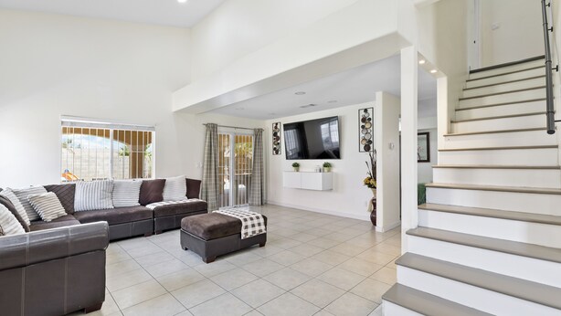 A spacious living room with a large sectional sofa, a small ottoman, a flat-screen TV mounted on the wall, and a staircase leading to an upper floor. - Patio w/ Grill + Pool Table , In-Unit Laundry (North Las Vegas)