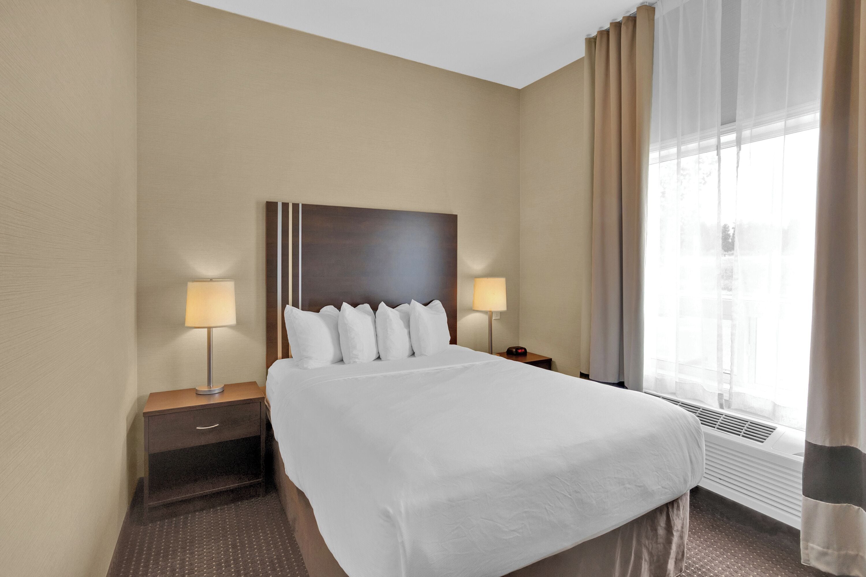 A hotel room with a large bed, two bedside tables with lamps, a headboard, and a window with curtains.