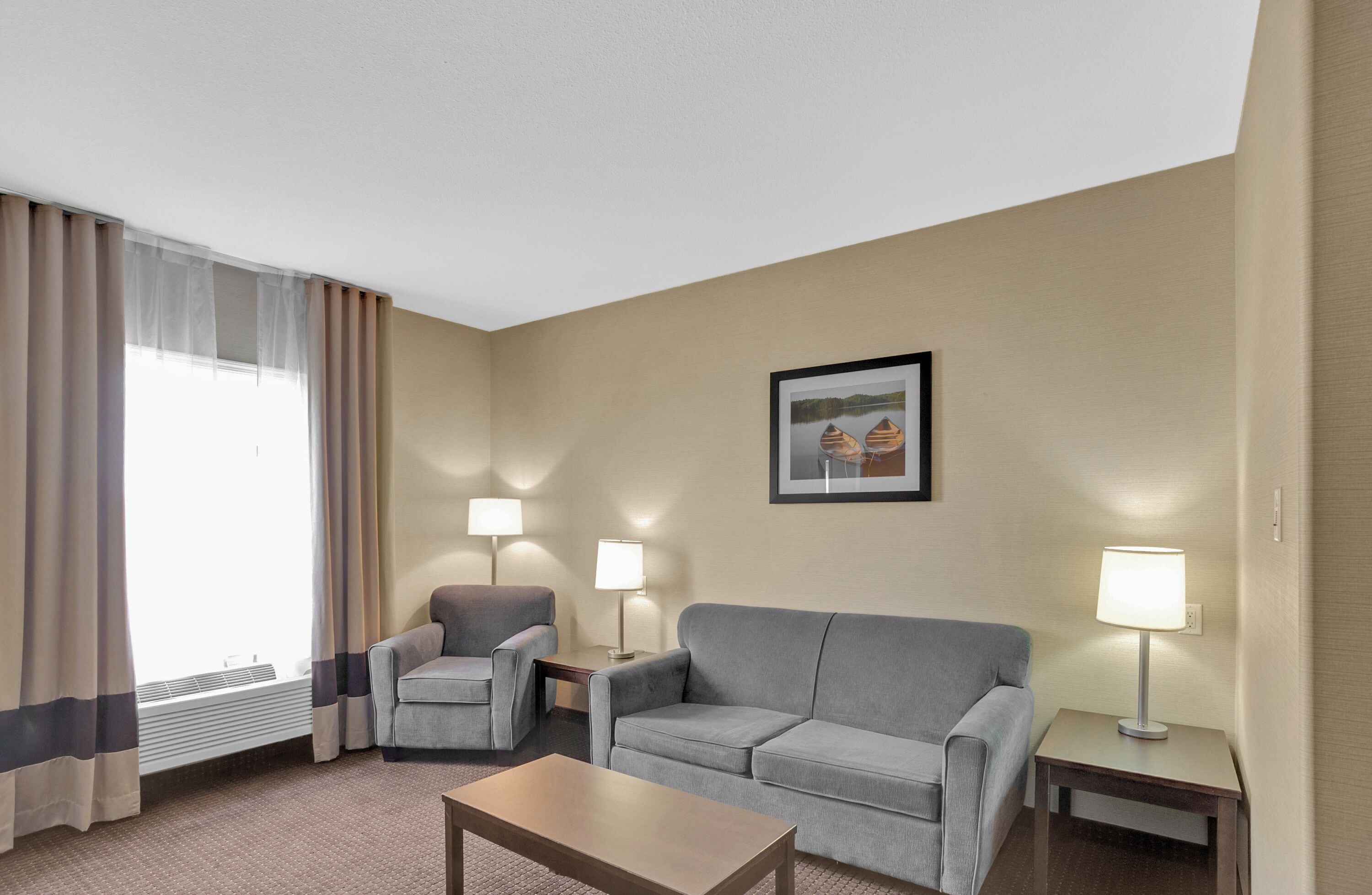 Habitación de hotel con un sofá gris, un sillón más pequeño, una mesa de centro de madera, dos lámparas de noche y un cuadro en la pared.
