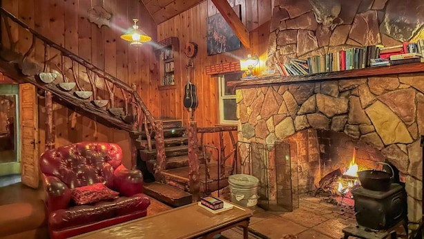 L’intérieur d’un chalet accueillant, avec une cheminée en pierre, un canapé en cuir rouge, un escalier en bois et une bibliothèque remplie de livres.