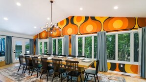 A dining area with a large table set for six, surrounded by chairs, large windows with curtains, and a modern chandelier.