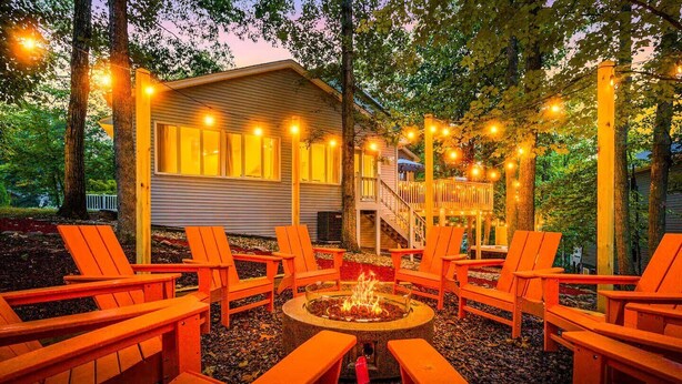 A cozy outdoor seating area with wooden chairs and a fire pit, surrounded by trees and string lights.
