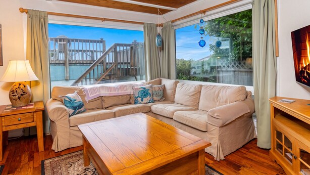 Een gezellige woonkamer met een houten plafond, een beige hoekbank, een houten salontafel en een groot raam met uitzicht op een houten terras en water.