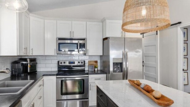 A modern kitchen with white cabinets, stainless steel appliances, a large island, and a woven pendant light. - Charming 3-bedroom cottage in Coral with WiFi and AC (Coral)