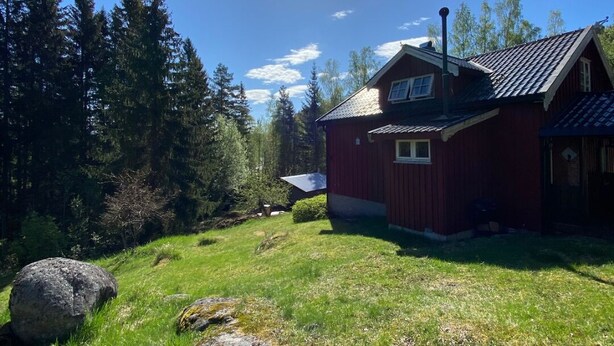 Une maison en bois rouge, avec un toit en pente, entourée d’une cour verdoyante et de nombreux arbres.