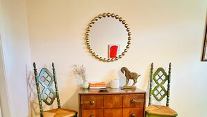 A room with a wooden floor, a wooden cabinet with drawers, two chairs with intricate designs, a mirror with a beaded frame, and a red abstract painting on the wall. - Charming 2-bedroom bungalow in welcoming Austin with WiFi, AC (Austin)