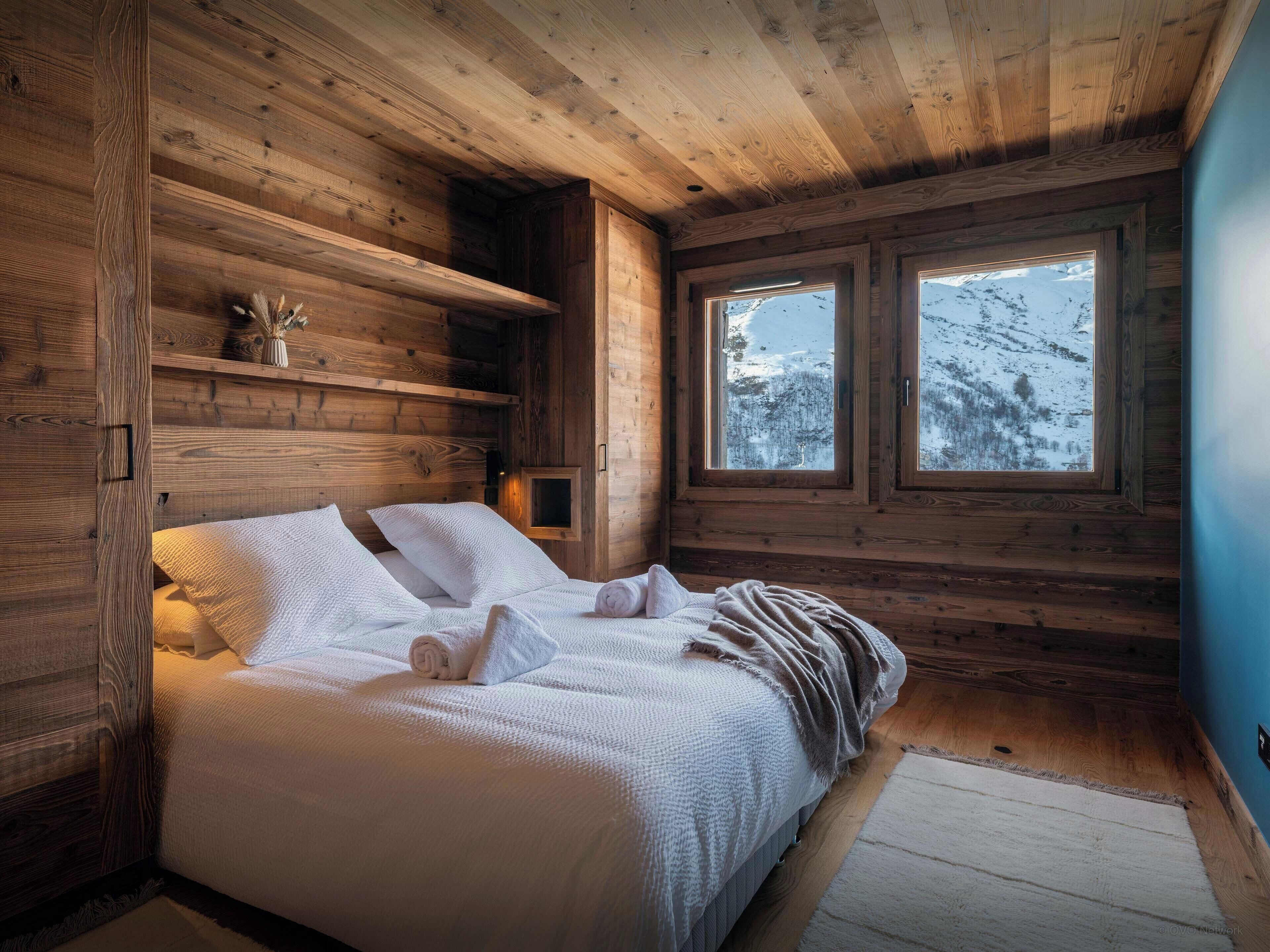 A cozy wooden cabin bedroom with a bed, a window with a snowy view, and a shelf with decorative items.