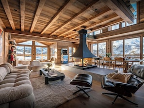 Amplia sala de estar con chimenea, vigas de madera y ventanas grandes que ofrecen vistas a un paisaje nevado.