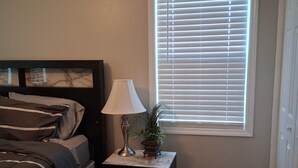 A bedroom with a bed, a nightstand with a lamp, a potted plant, and a window with blinds. - ABM Southern Vacation Rental 
3-bedroom house in marvelous Savannah with WiFi (Savannah)