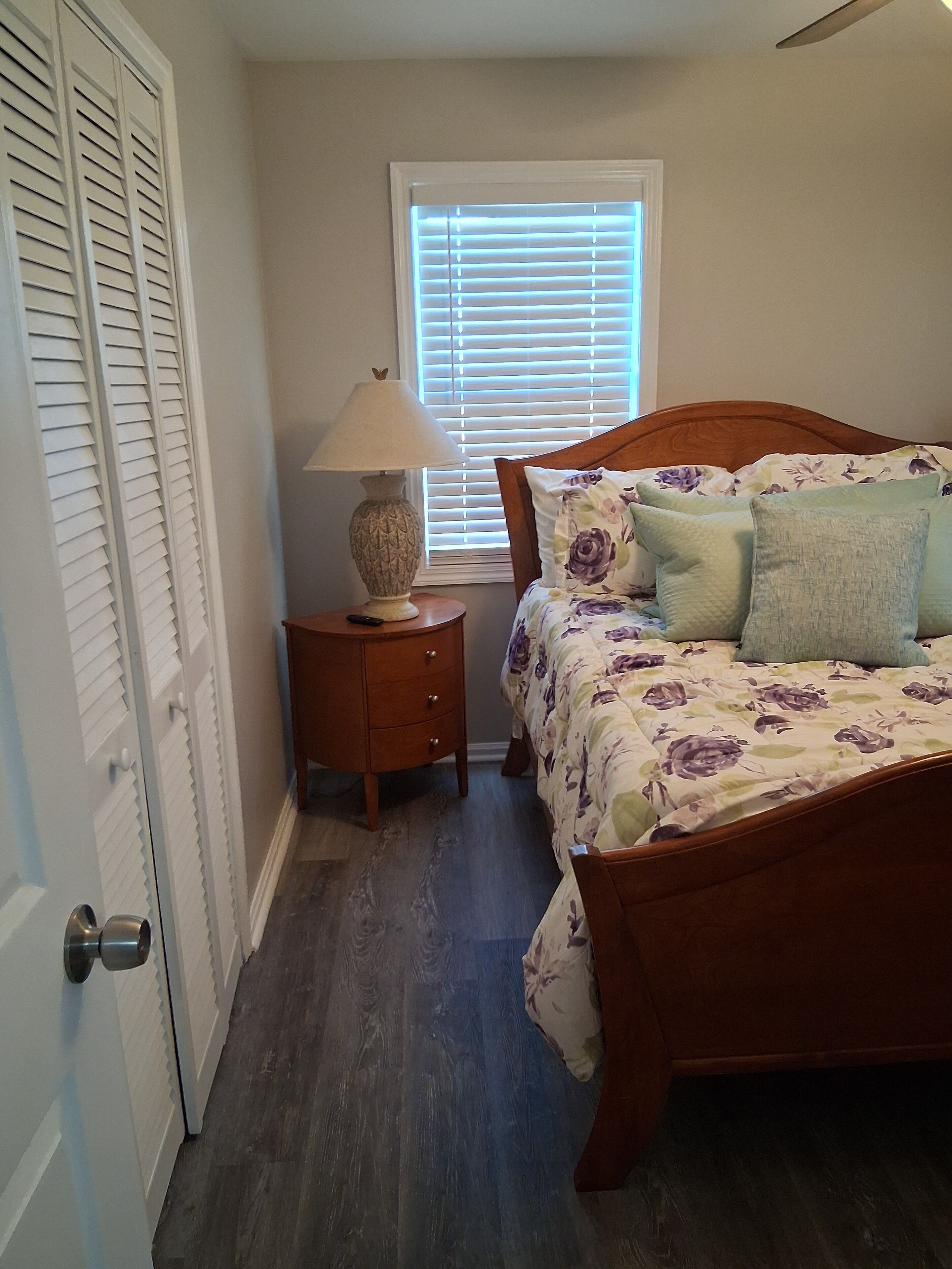 A bedroom with a wooden bed, a nightstand with a lamp, a window with blinds, and a door with louvered shutters.