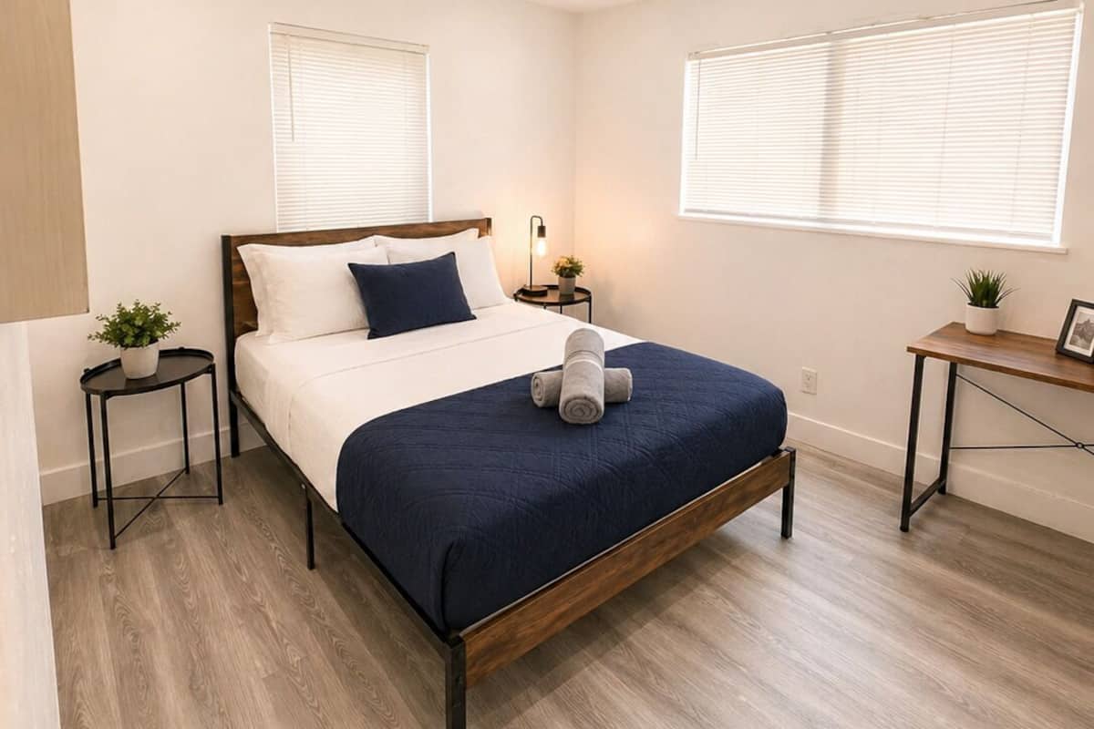 Une chambre à coucher comprenant un lit, deux tables de chevet, une commode, un petit bureau et deux fenêtres équipées de stores.