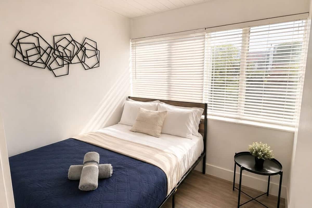 Une chambre à coucher avec un lit, une table de chevet agrémentée d’une plante, une fenêtre avec des stores et une œuvre d’art géométrique accrochée au mur.