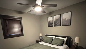 Een slaapkamer met een bed, een plafondventilator, een raam met zonwering en drie ingelijste foto's aan de muur.