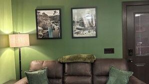 Een bruine leren bank met groene kussens, tegen een groene muur, met aan weerszijden twee zwart-wit foto's in lijsten.