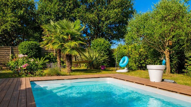 A rectangular swimming pool with a wooden deck surrounding it, set in a lush garden with various trees and plants. - Studio for 3 people – Shared pool (Les Sables-d´Olonne)