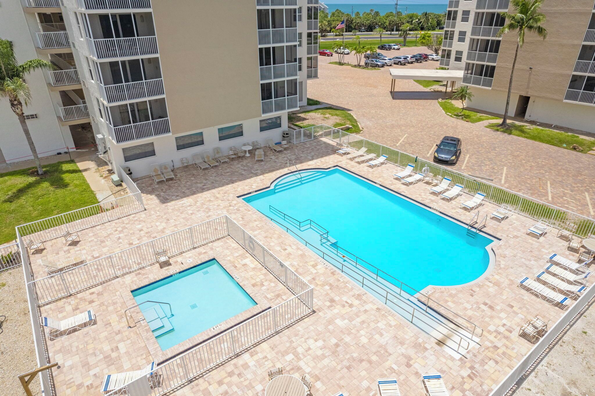 A rooftop pool area with multiple lounge chairs and a smaller pool, surrounded by a building with balconies and a view of the ocean.