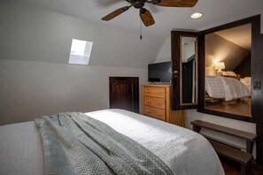 Un dormitorio con cama, cómoda de madera, ventilador de techo, televisión y un espejo grande que refleja la habitación.