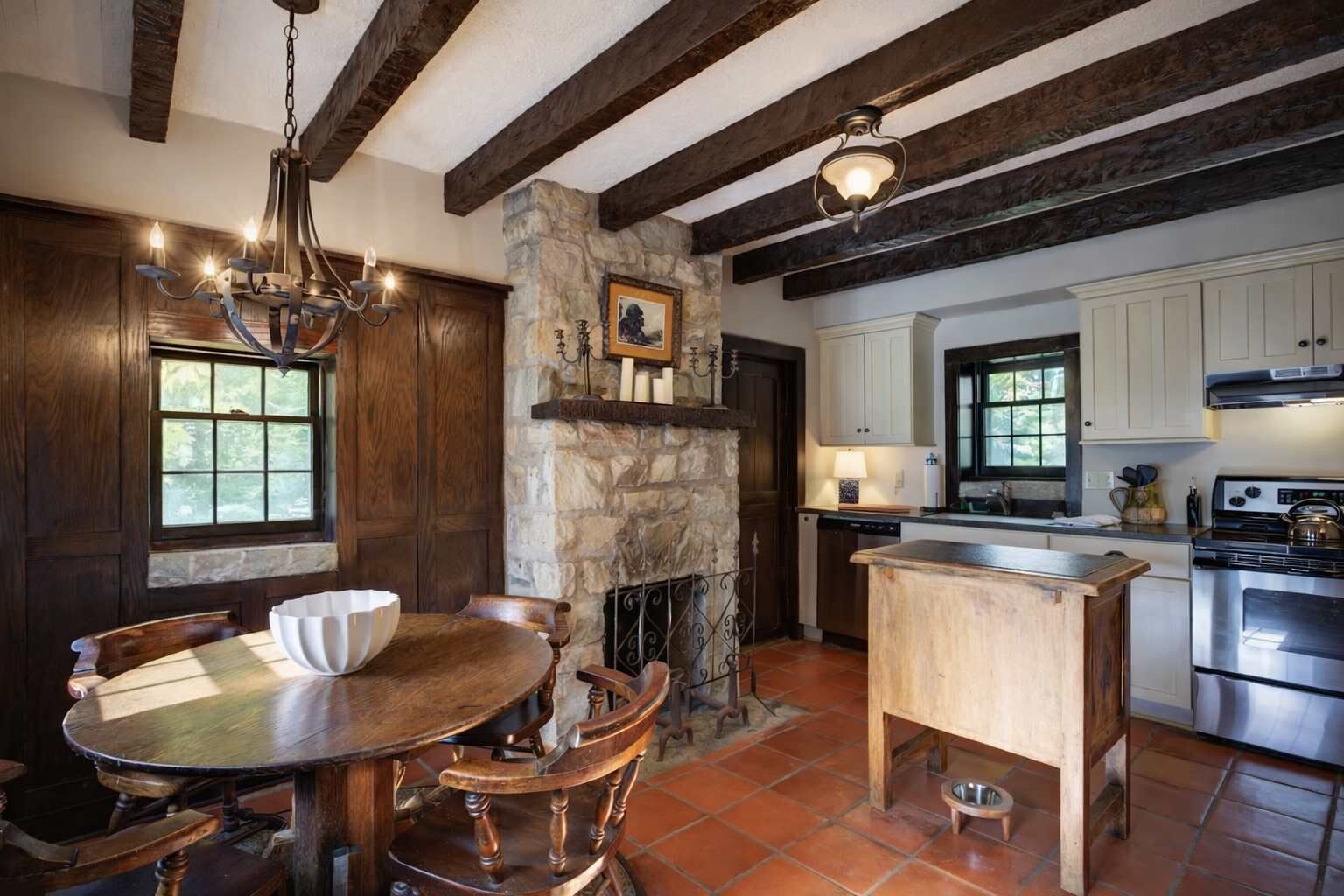 A rustic kitchen with a stone fireplace, wooden beams, and a dining area with a round table and chairs.